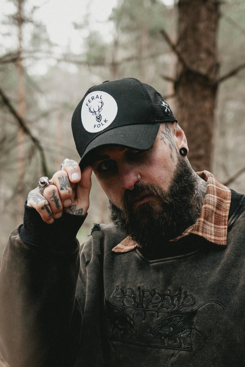 Man wearing a black Feral Folk cap with a logo, tattoos, and a plaid shirt in a forest setting.