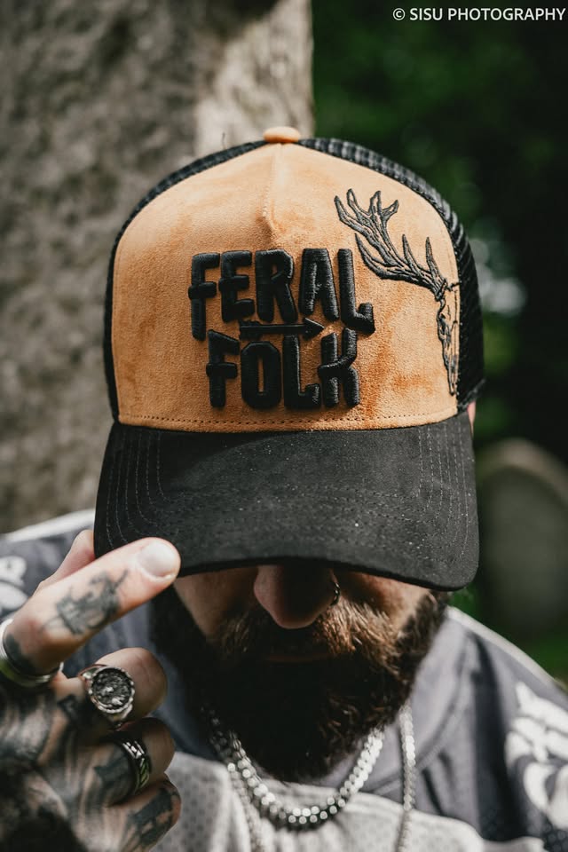 Person wearing a cap with 'Feral Folk' text and deer design, outdoors.