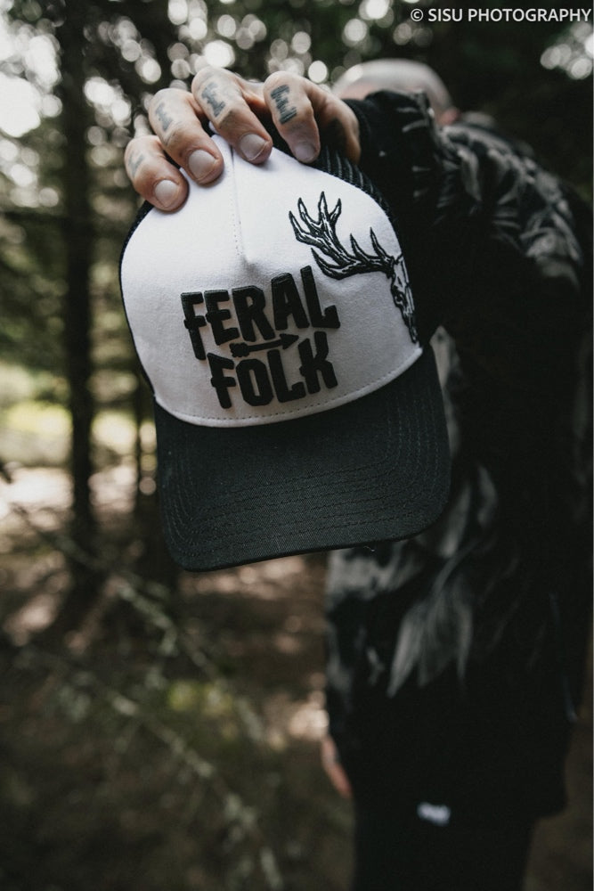Person holding a cap with 'Feral Folk' and deer emblem design in a forest setting.